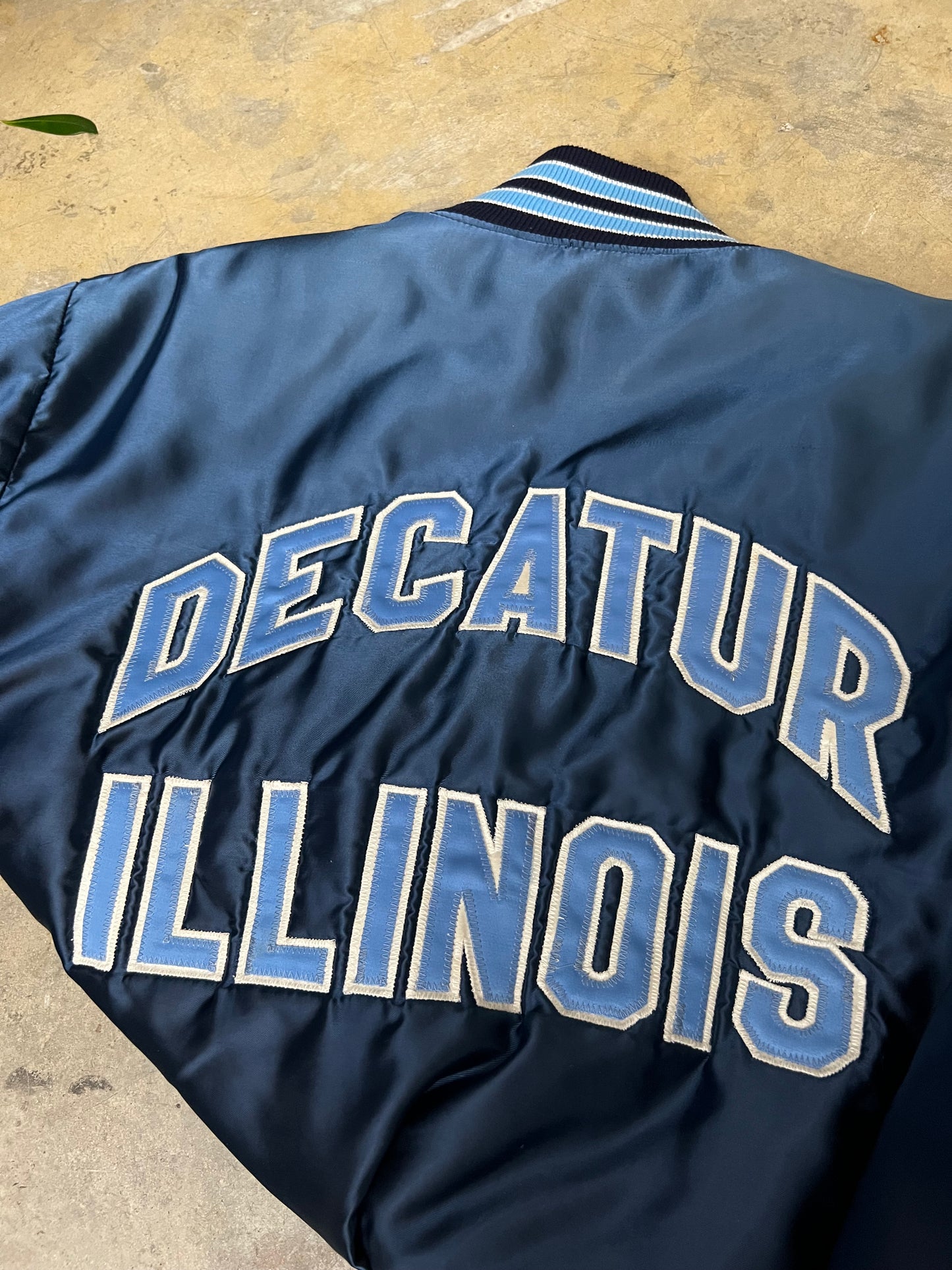 Vintage blue satin varsity bomber jacket features "Decatur Illinois" embroidered on the back in a light blue and white block font Large